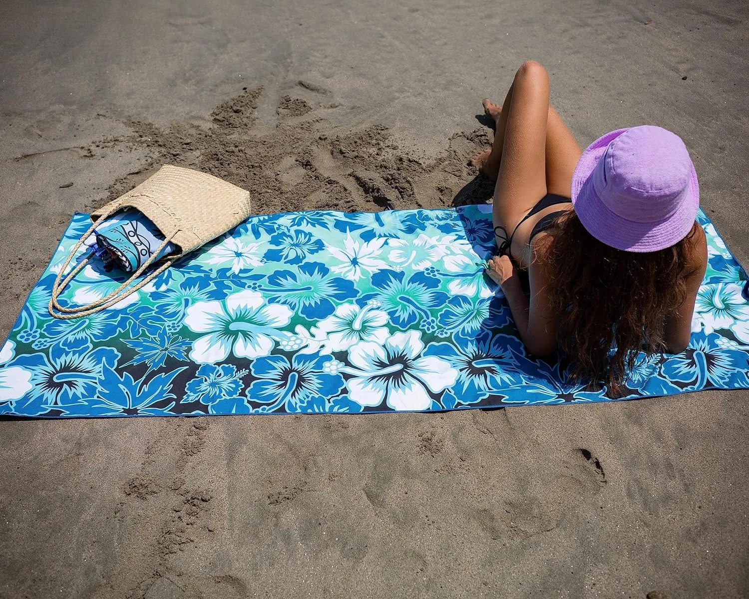 Blue and white floral beach towel spread on sand, with a person reclining on it and a straw bag alongside.