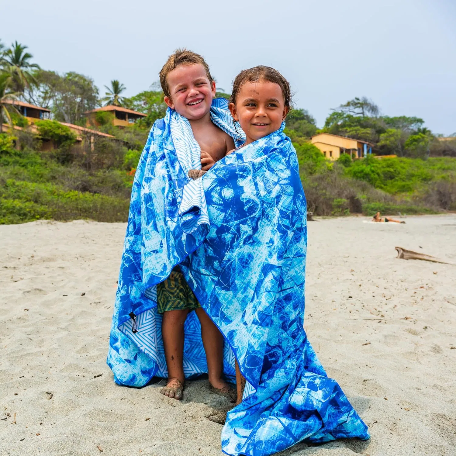 Two children wrapped in blue and white patterned microfiber beach towels on a sandy beach.
