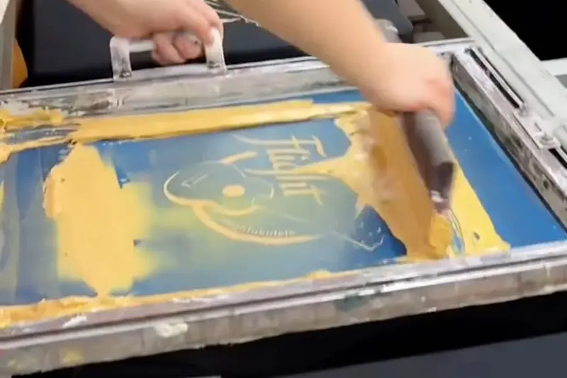 A worker pulling yellow ink across a screen with a squeegee to print a design.