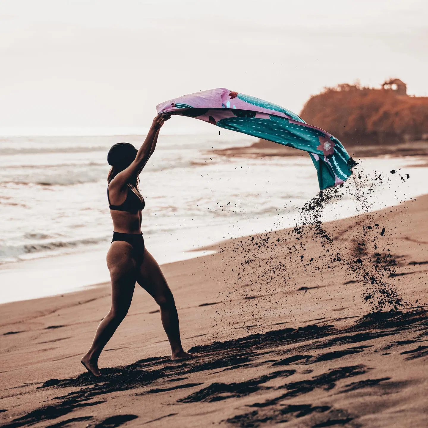 A person shaking sand off a pink and teal patterned beach towel beside the sea.