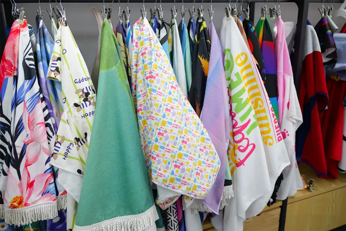 Printed and solid-colored beach towel samples hanging from metal clips on a display rail.