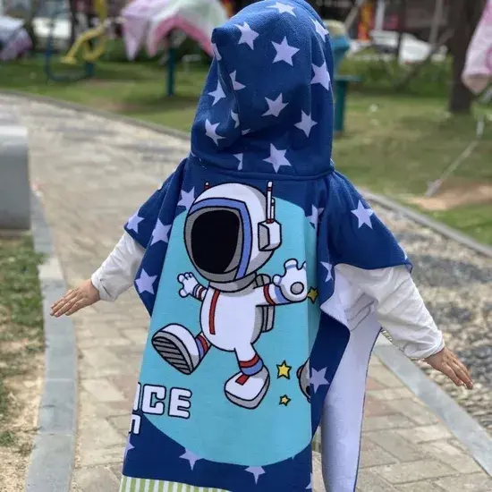 Child wearing a navy hooded poncho towel with white stars and a large astronaut illustration on a turquoise panel outdoors.