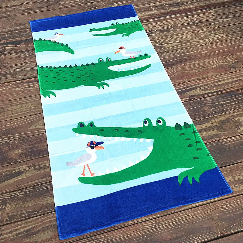 Beach towel with green cartoon crocodiles, pale blue stripes and dark blue end bands on a wooden floor.