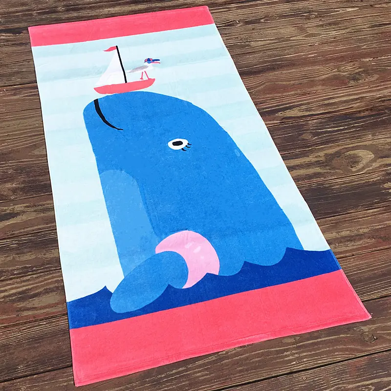 Beach towel with a blue cartoon whale, a small sailboat and pink end bands on a wooden floor.