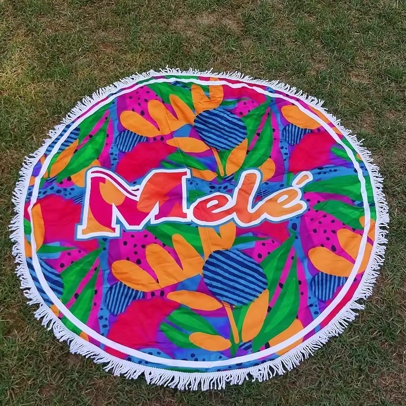 Round beach towel on grass with bright pink, orange, green and blue patterns, large central lettering and white fringe.