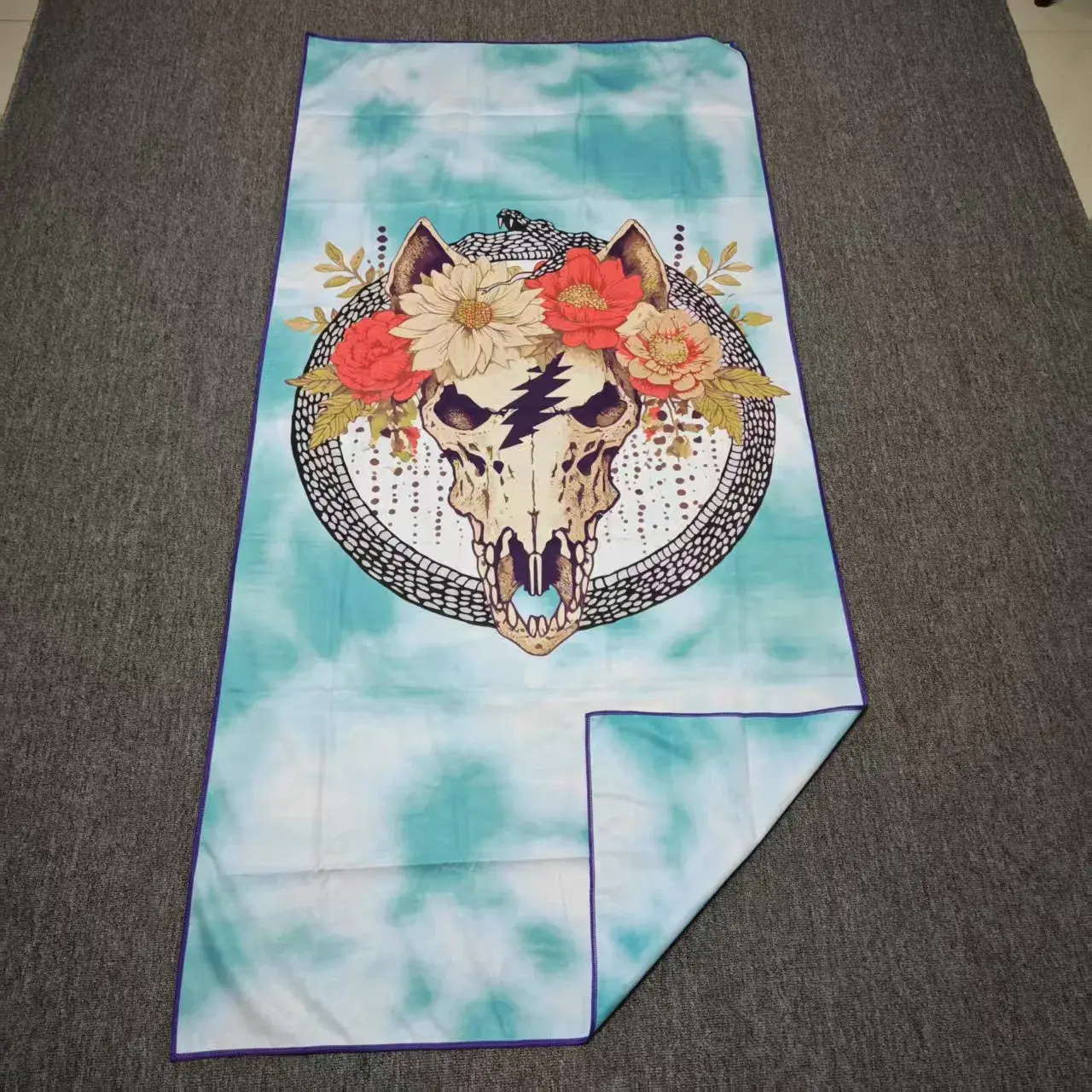 Aqua and white beach towel with an animal skull, red and cream flowers and a circular snake motif on gray carpet.