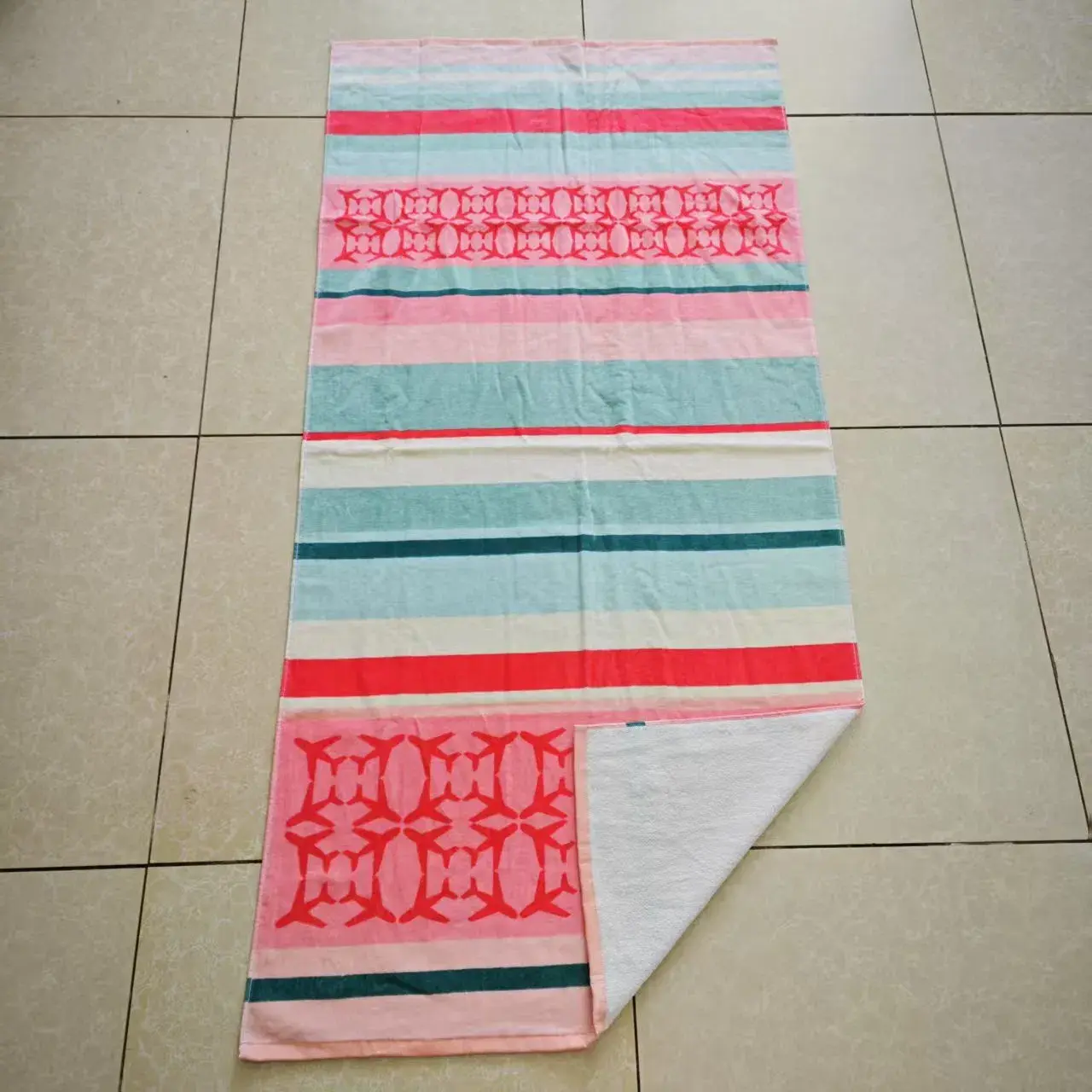 Pink and aqua striped beach towel with red geometric motifs and a folded corner showing its white reverse.