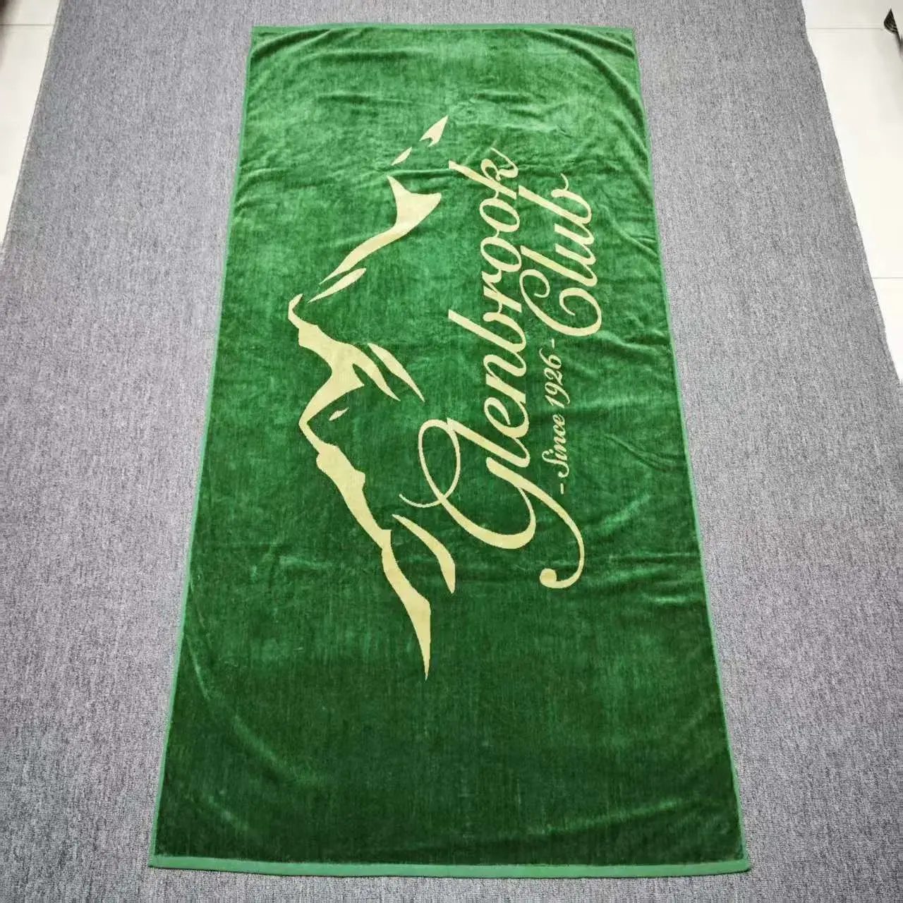Green beach towel with a cream mountain outline and script club lettering laid flat on gray carpet.
