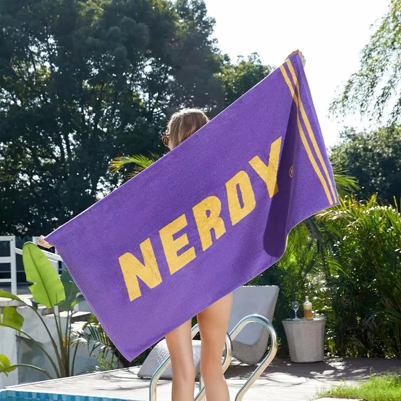 Purple beach towel with large yellow lettering and yellow border stripes held open beside a pool.