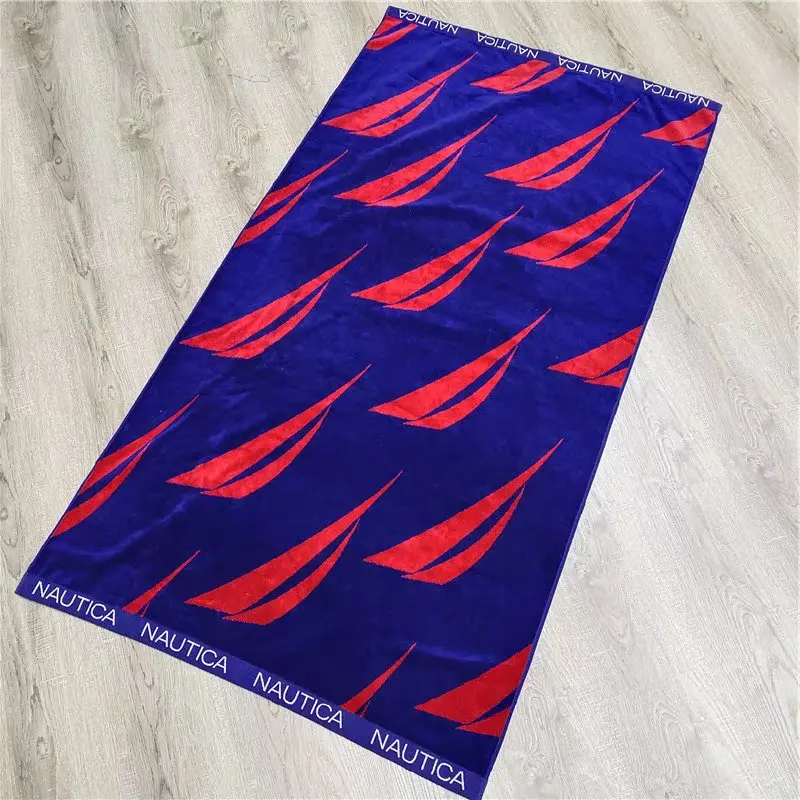 Blue beach towel with red sail motifs and white lettering along the ends on a light wood floor.