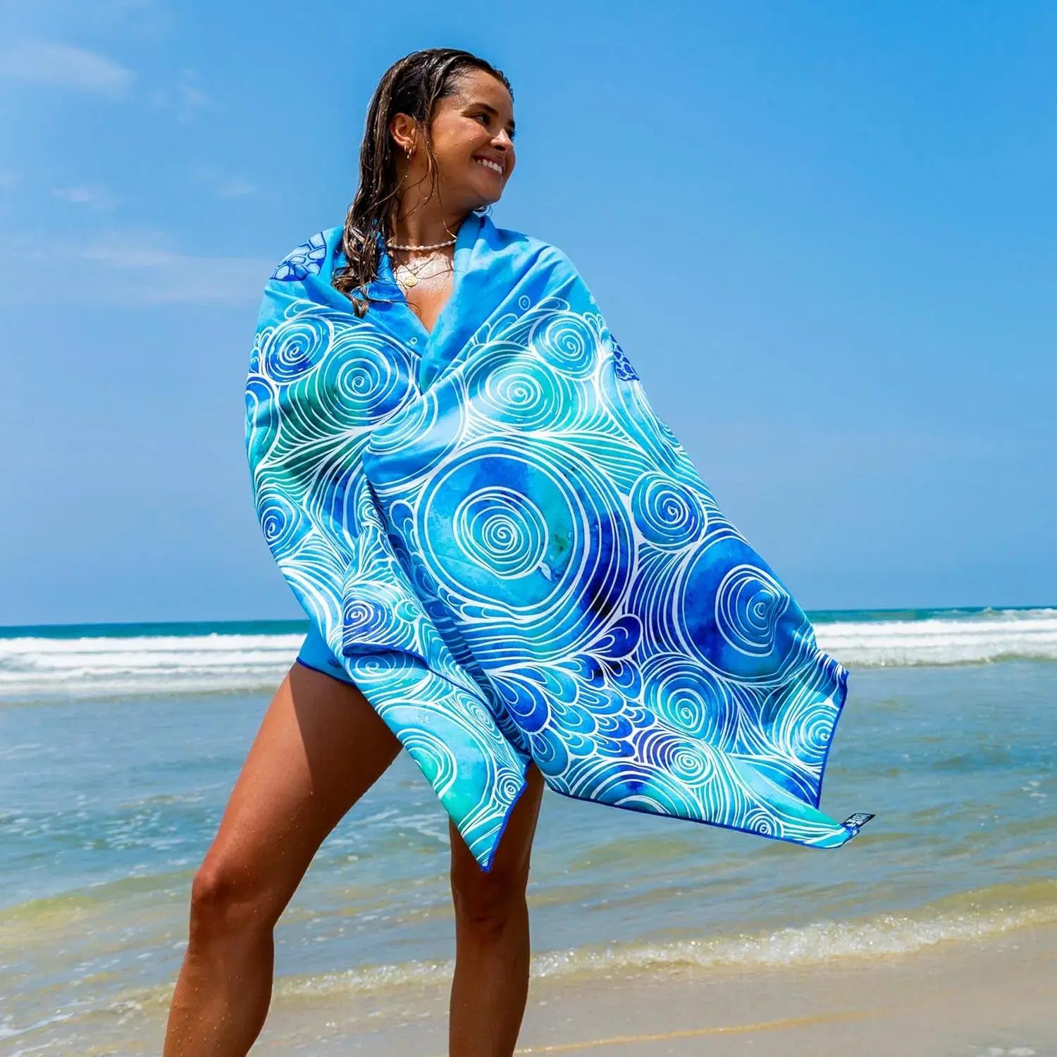 Blue and turquoise beach towel with white swirling wave patterns wrapped around a woman at the shoreline.
