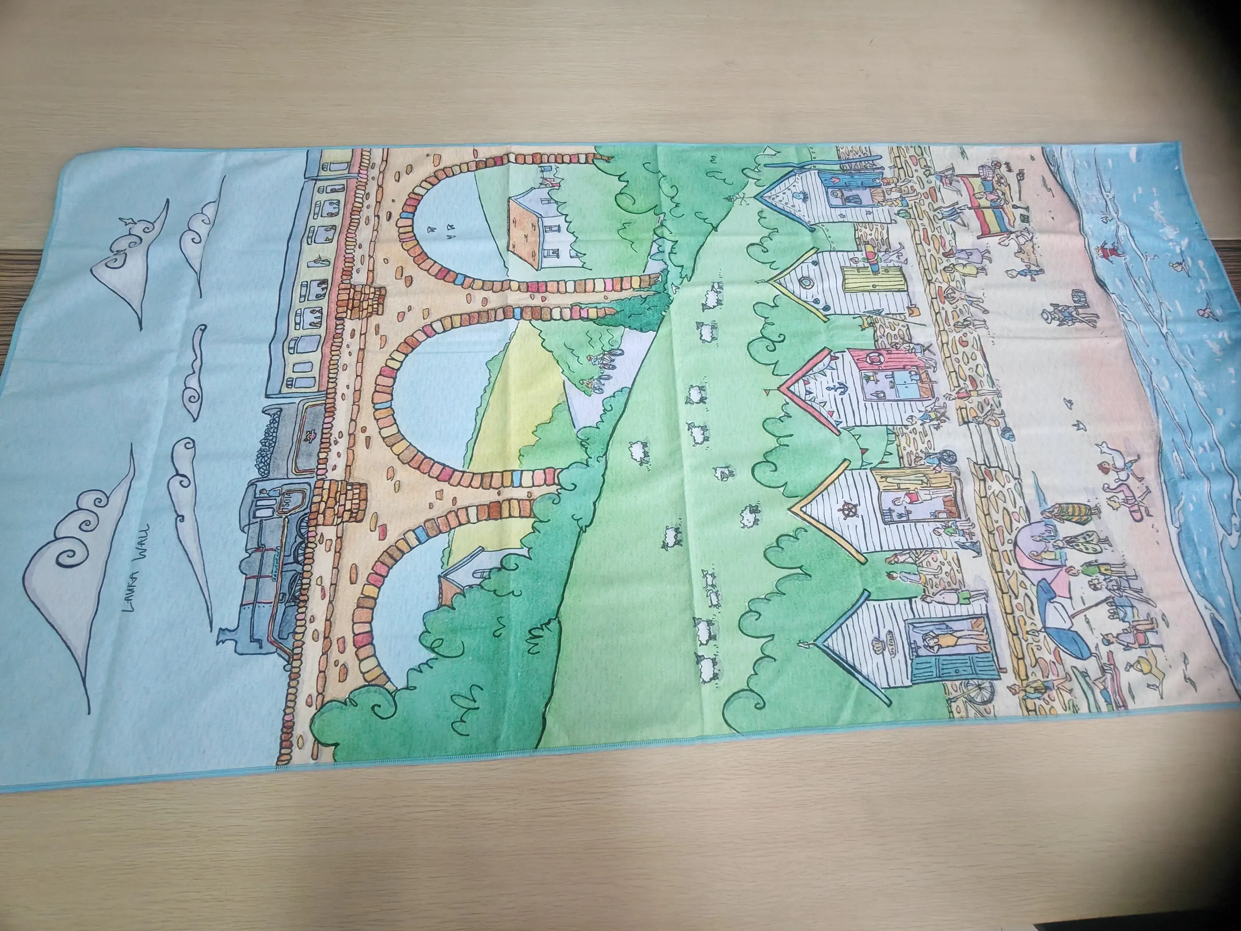 Beach towel with a seaside illustration of a train on an arched viaduct, green countryside, beach huts and people by the sea.
