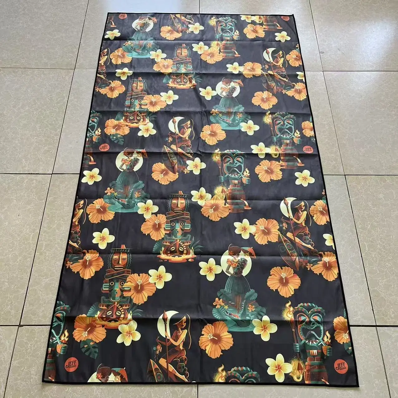 Dark beach towel with colorful tiki figures, illustrated dancers, orange hibiscus flowers and pale yellow flowers.