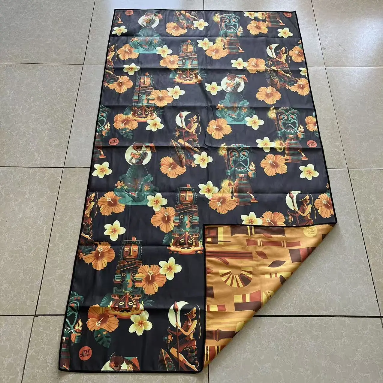 Tiki and floral beach towel with a corner folded back to show a golden brown reverse with geometric patterns and black stitched edging.