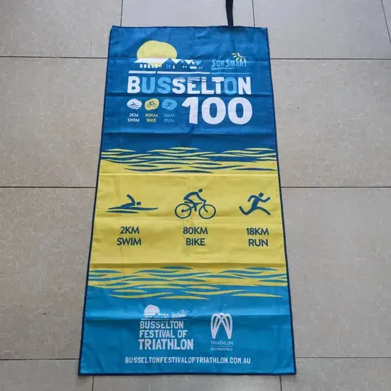 Blue and yellow triathlon event beach towel with swimming, cycling and running icons laid flat on a tiled floor.
