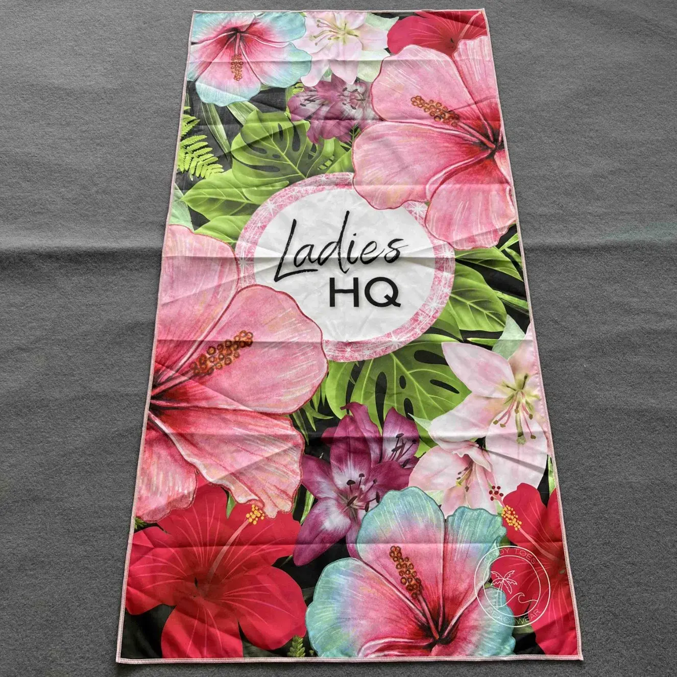 Beach towel with large pink and red flowers, green tropical leaves and a central white circle with black lettering.