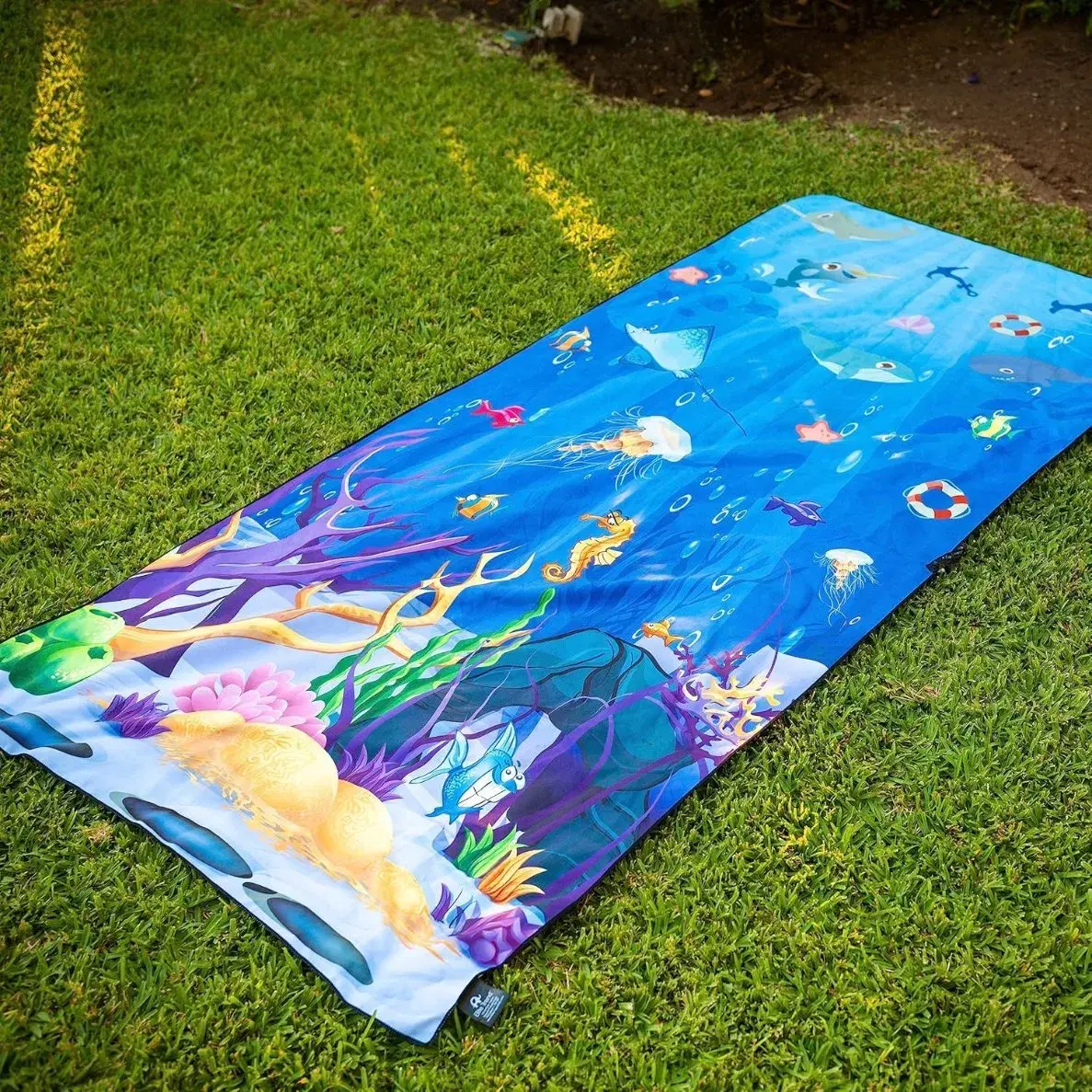 Blue beach towel with fish, jellyfish, seahorses and colorful coral illustrations laid out on grass.