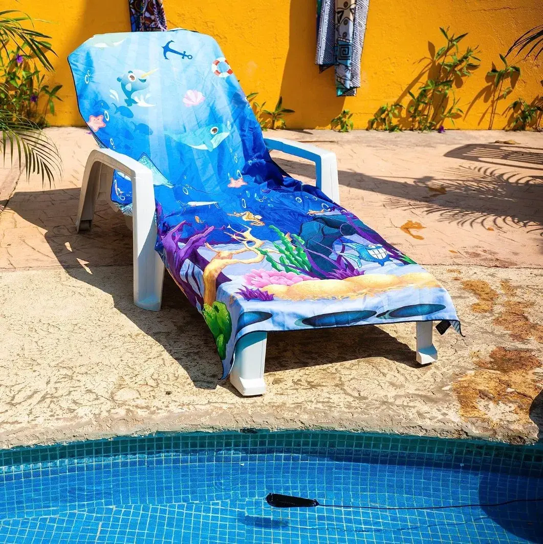 Blue underwater-scene beach towel draped over a white lounger beside a swimming pool.