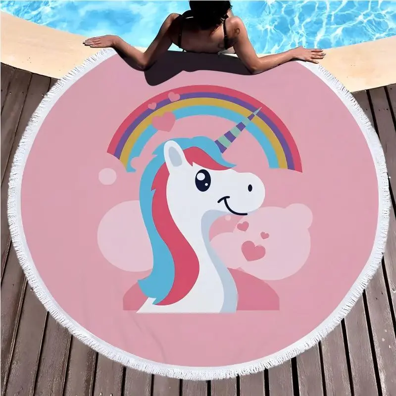 Pink round beach towel with a unicorn and rainbow print and white fringe beside a swimming pool.