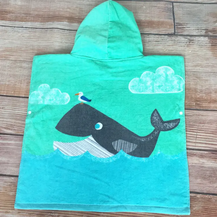 Turquoise hooded poncho towel with a dark whale, a small seabird, white clouds and blue waves laid flat on a wood-patterned surface.