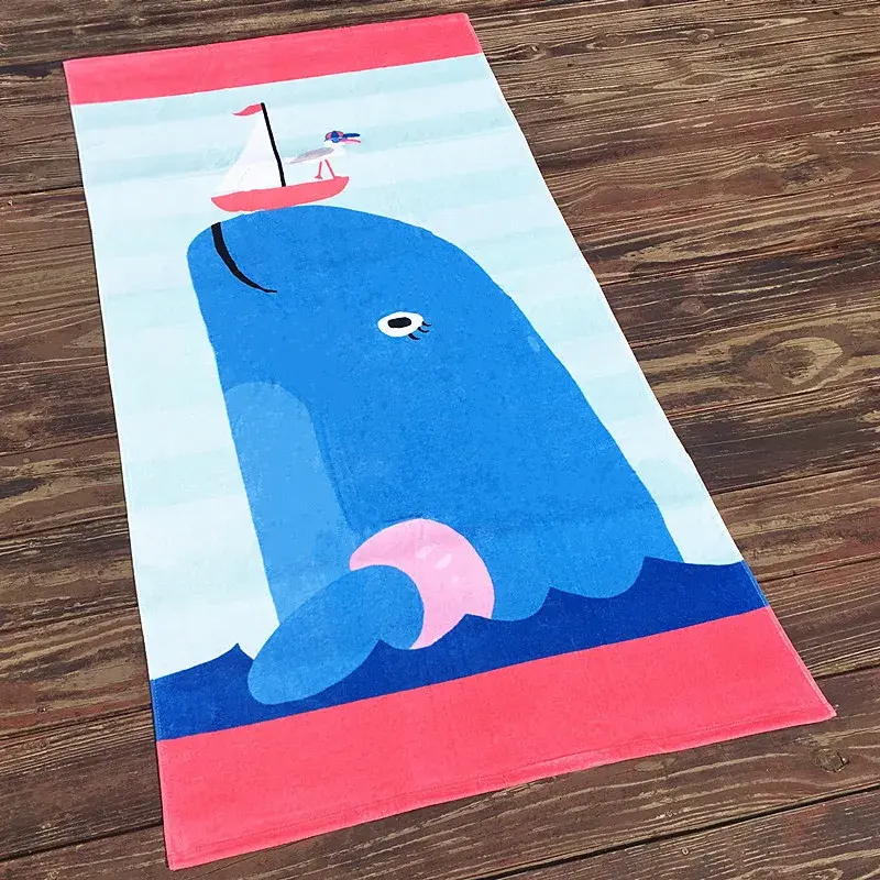 Beach towel with a blue cartoon whale, a small sailboat and pink end bands on a wooden floor.