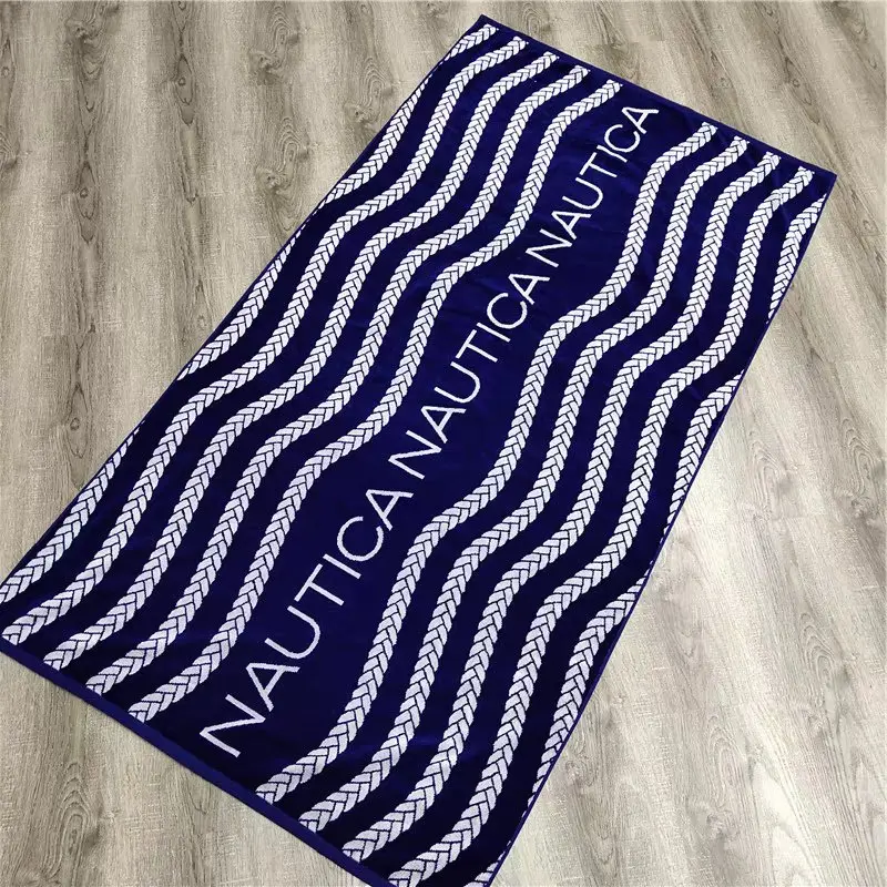 Navy beach towel with white wavy rope motifs and large white lettering on a light wood floor.