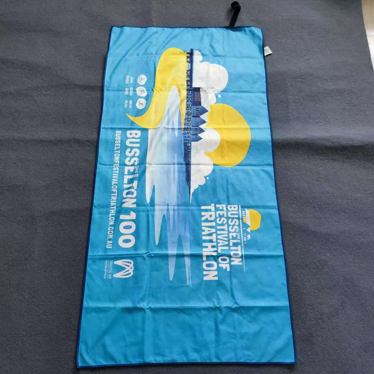 Blue microfiber beach towel with a yellow and white event print laid flat on a gray surface.