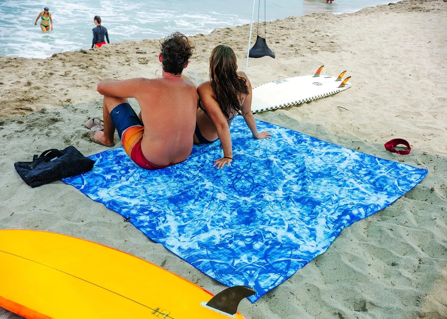 Two people sitting on an oversized blue printed beach towel beside surfboards.