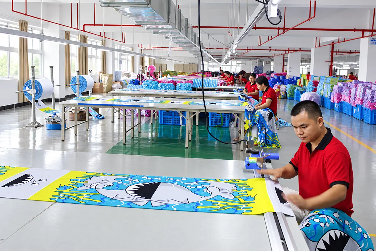 Workers handling shark-print beach towel fabric at long worktables in a production workshop.