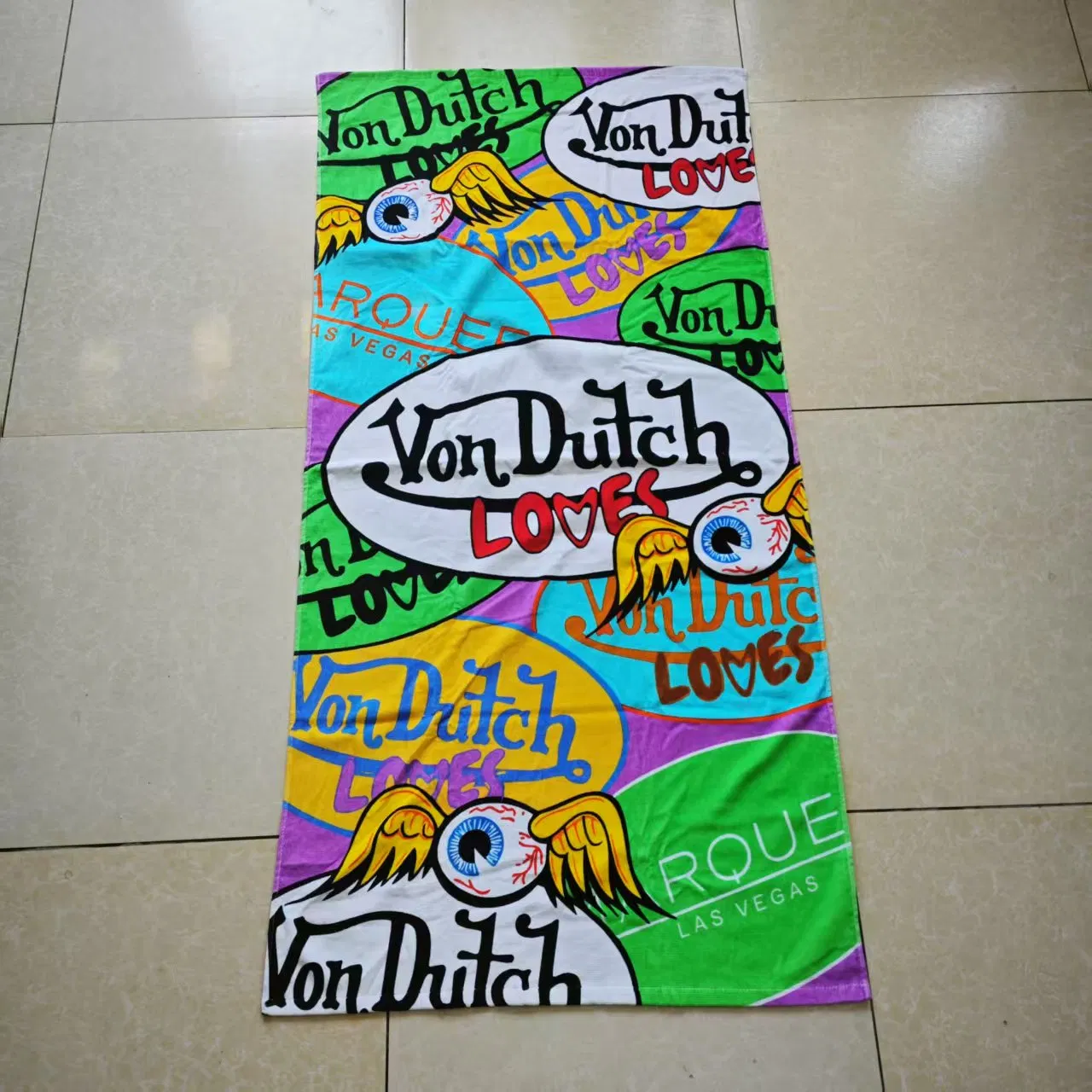 Colorful printed beach towel sample with bold lettering and winged-eye graphics laid flat on a tiled floor.