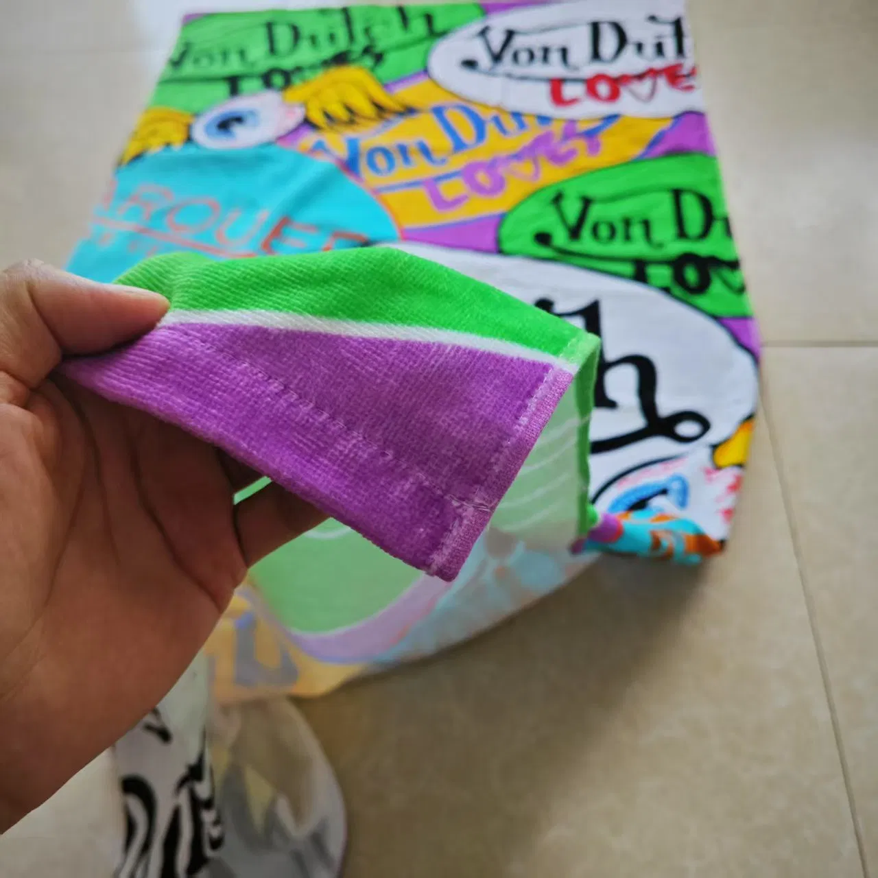 Hand holding the green and purple corner of a printed beach towel, showing the fabric texture and stitched hem.